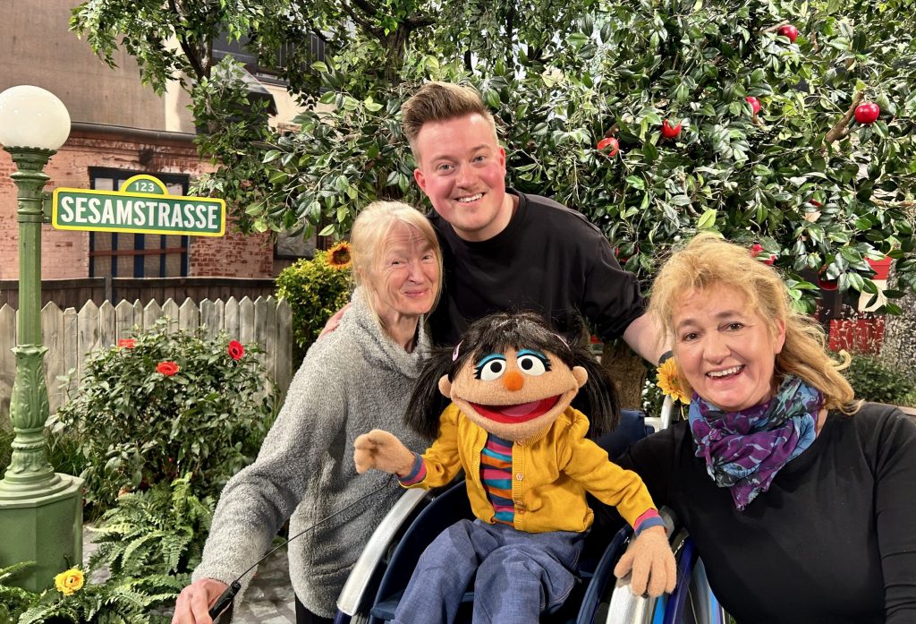 Die Puppenspielerinnen Iris Schleuss und Charlie Kaiser zusammen mit ihrer Puppe Elin und René Schaar. Elin trägt eine gelbe Strickjacke und nutzt einen Rollstuhl.