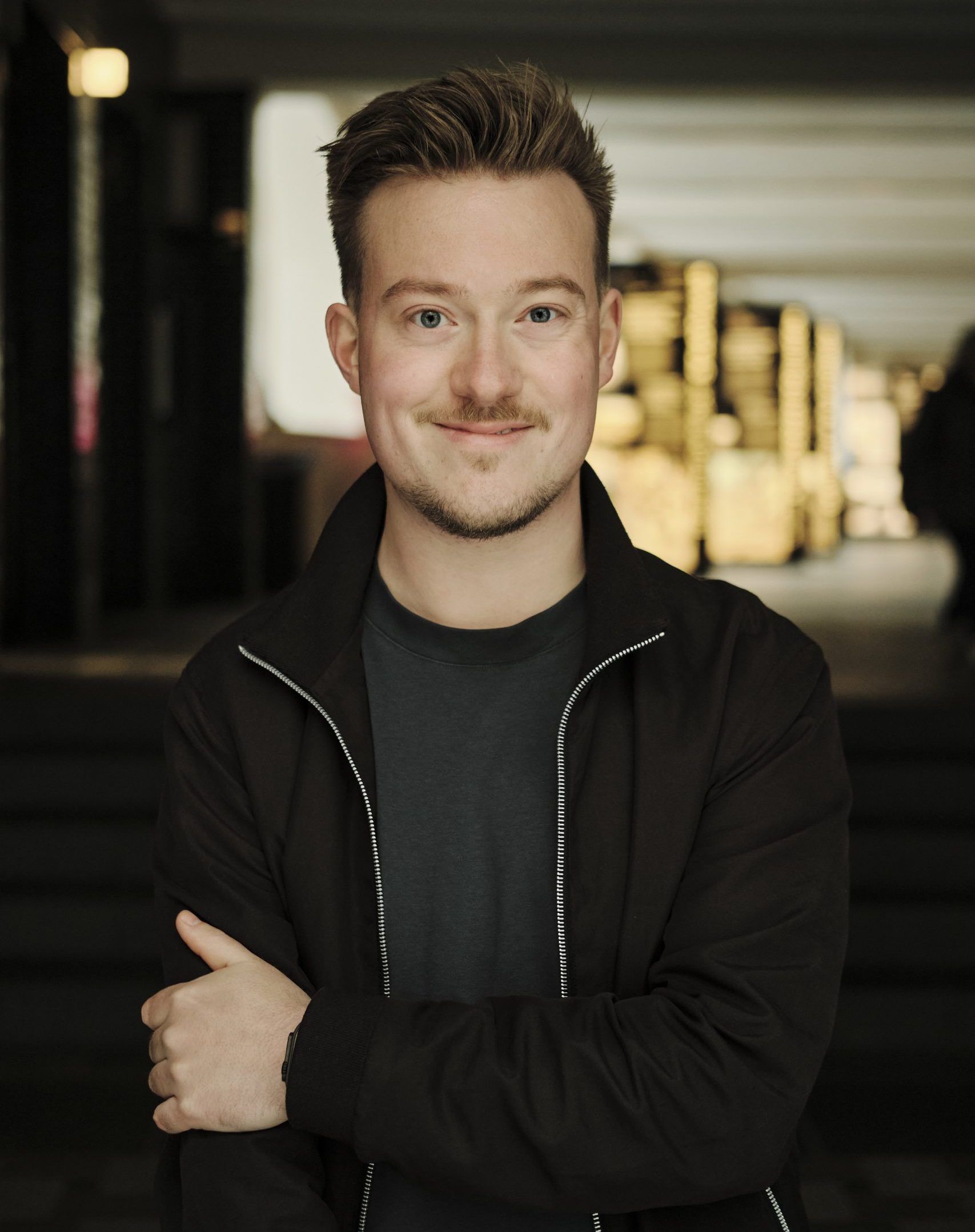 Portrait von René, schwarze Jacke, kurze blonde Haare, graues Shirt.